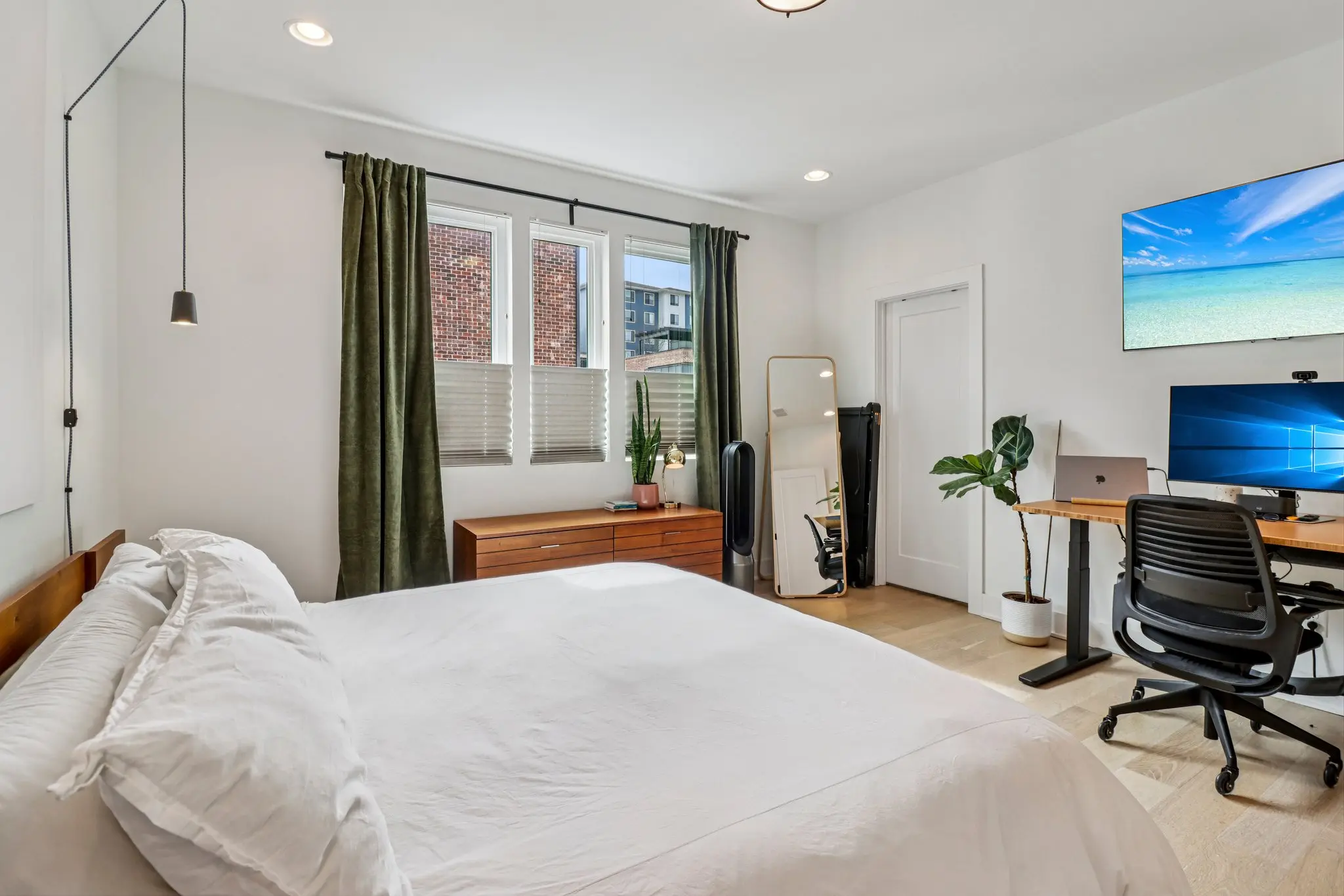 Bedroom set up with a standing desk and task chair alongside the bed, with green velvet drapes at the window.