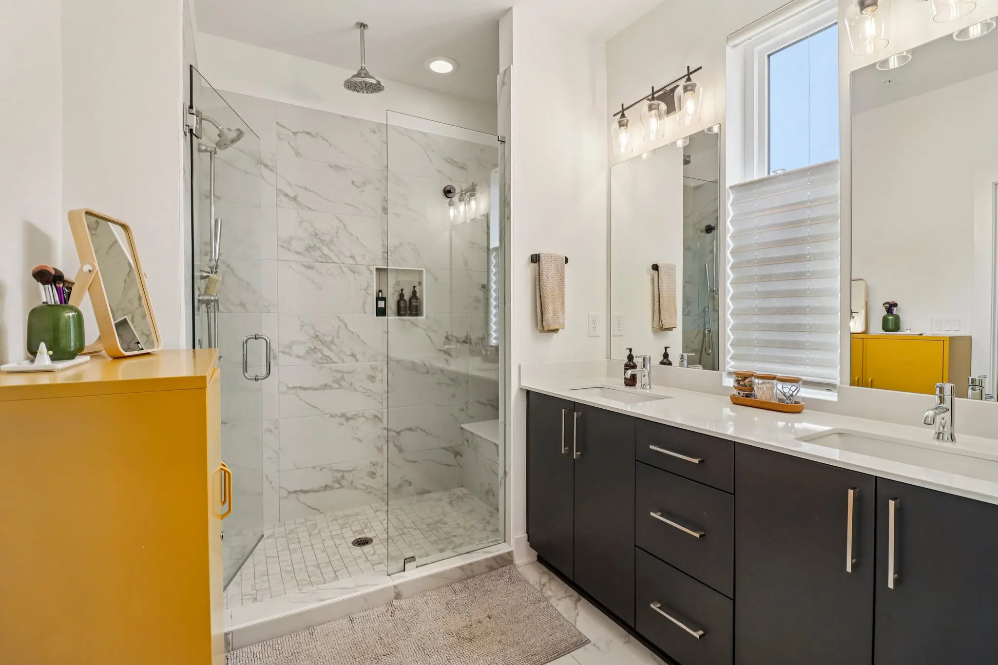 Primary bathroom with a glass-enclosed rain shower in marble-look tile, built-in bench, and a dark double vanity with quartz counter.